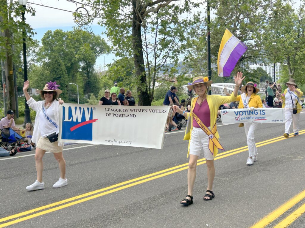 Members of the League of Women Voters march along the streets of Portland holding banners and waving to community as part of the annual Grand Floral Rose Parade.